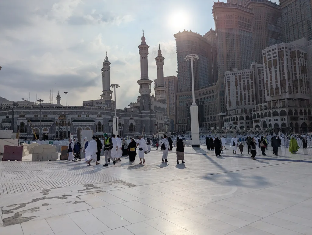 Pilgrims walking across the marble courtyard of the Grand Mosque in Makkah during the day, with the Abraj Al-Bait towers behind