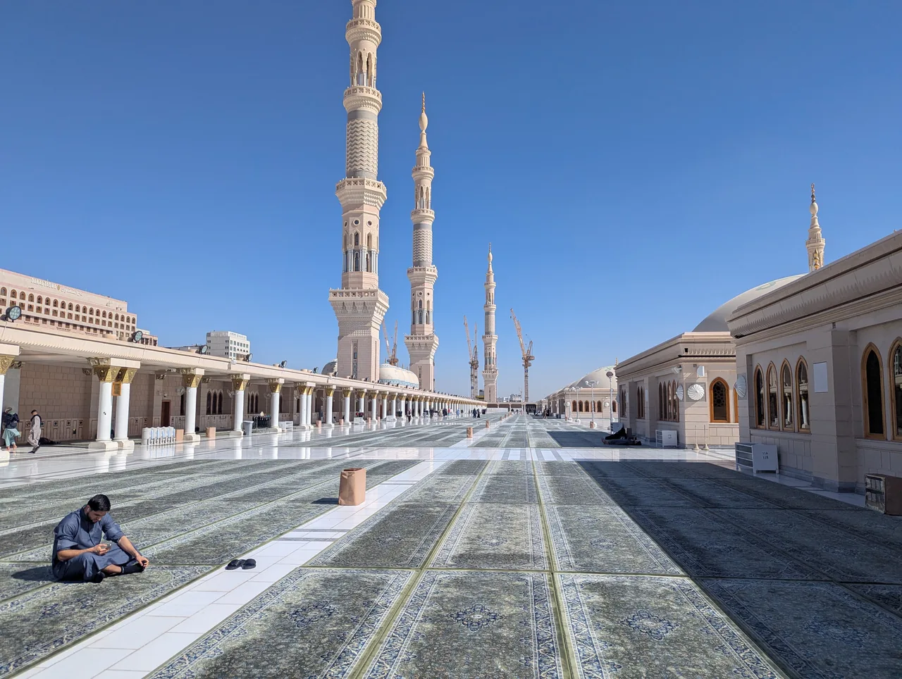 A worshipper resting on the patterned carpets in the vast courtyard of Al-Masjid an-Nabawi, the Prophet's Mosque in Madinah, with its minarets rising against a clear blue sky