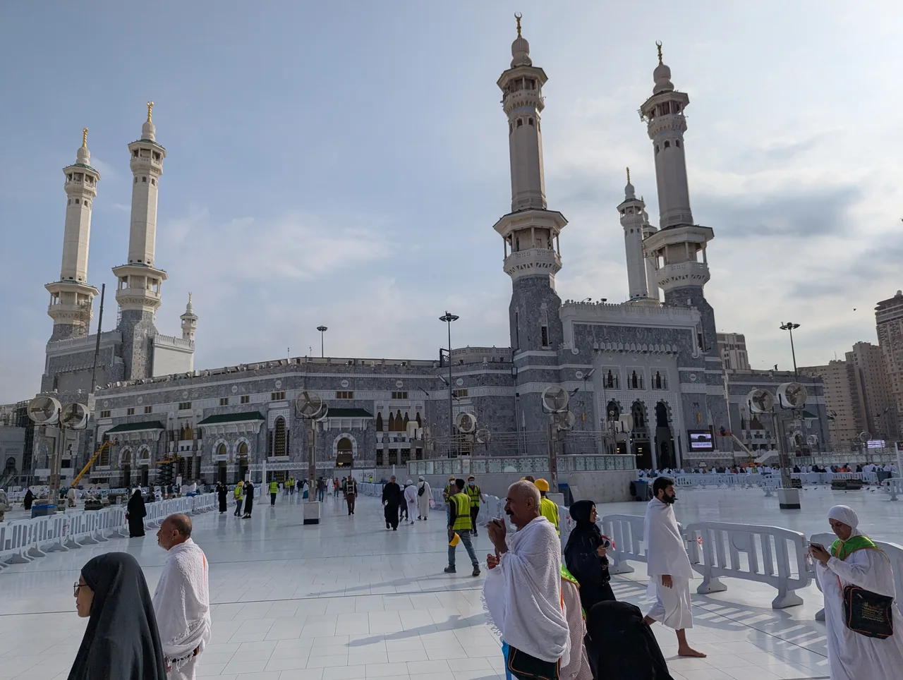Pilgrims in white ihram crossing the marble plaza outside Masjid al-Haram in Makkah, with the mosque's minarets rising under a daytime sky